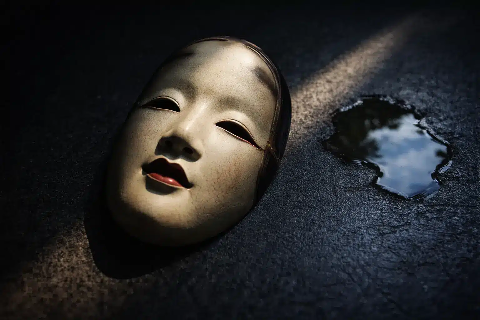 Eine japanische Theatermaske liegt abgelegt auf dunklem Boden neben einer Pfütze