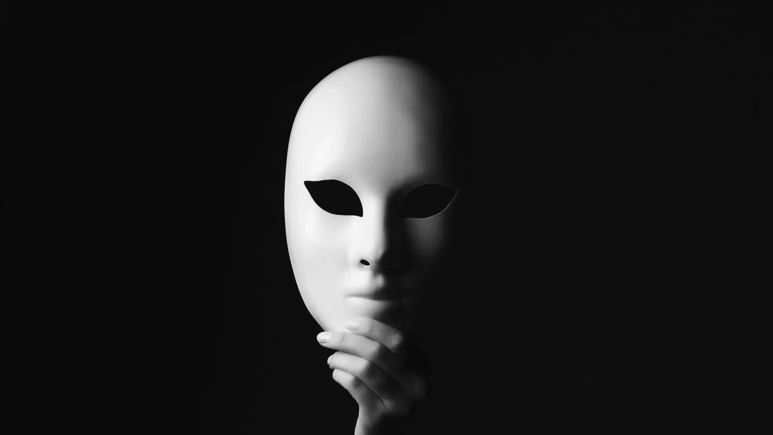 Weiße Maske vor dem Gesicht Eine Hand hält eine weiße Maske vor ein Gesicht, das im Dunkel verschwindet